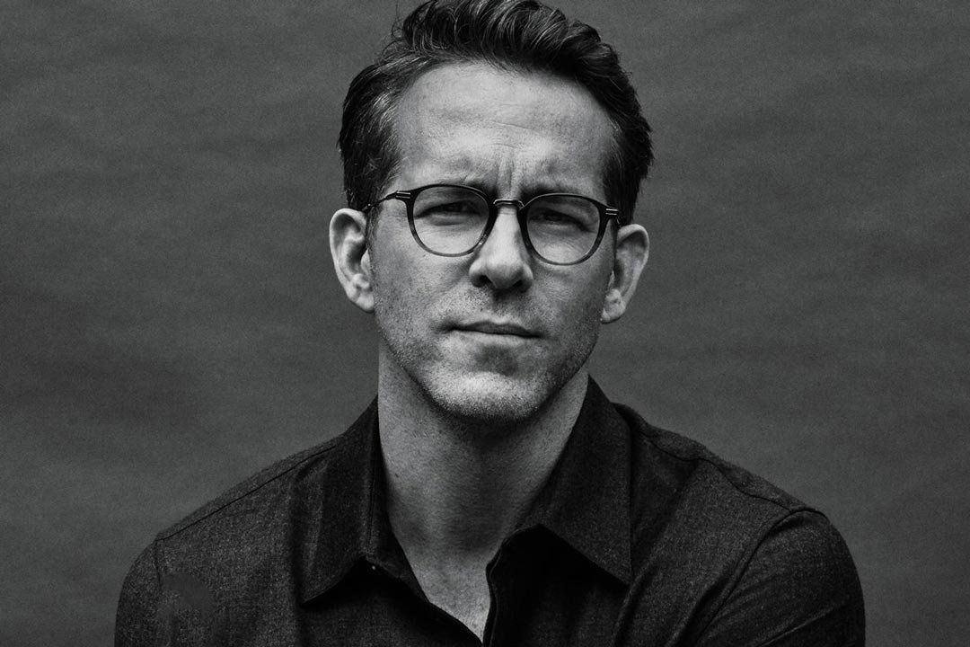 Greyscale image of man sitting in front of plain grey background wearing a shirt and oval spectacles
