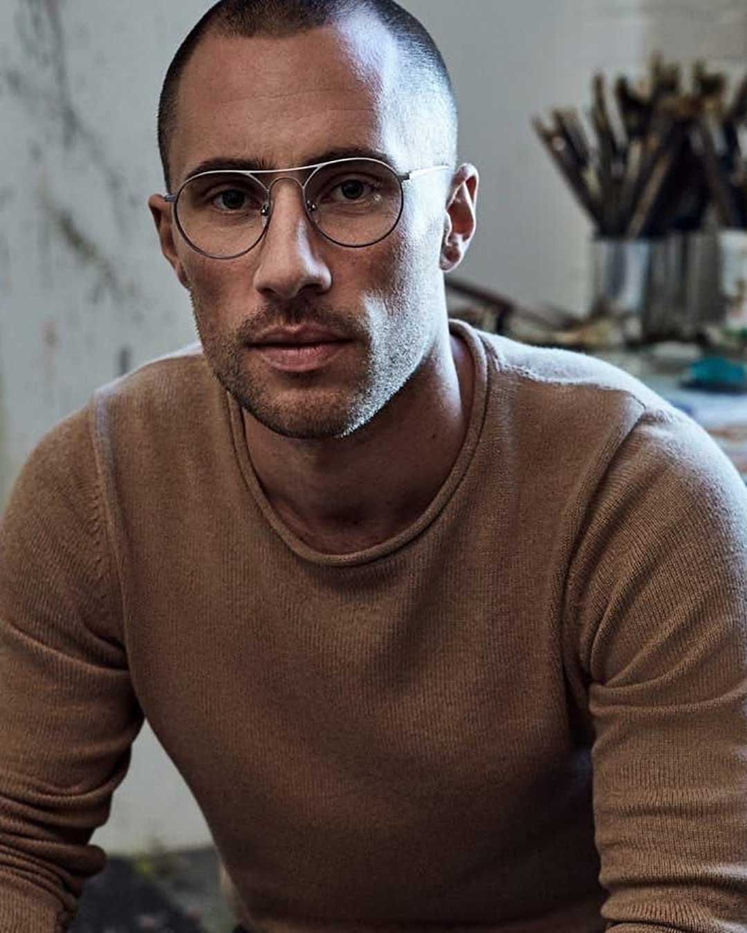 Man sitting in artist studio wearing cream jumper and wire rimmed Pilot glasses frame
