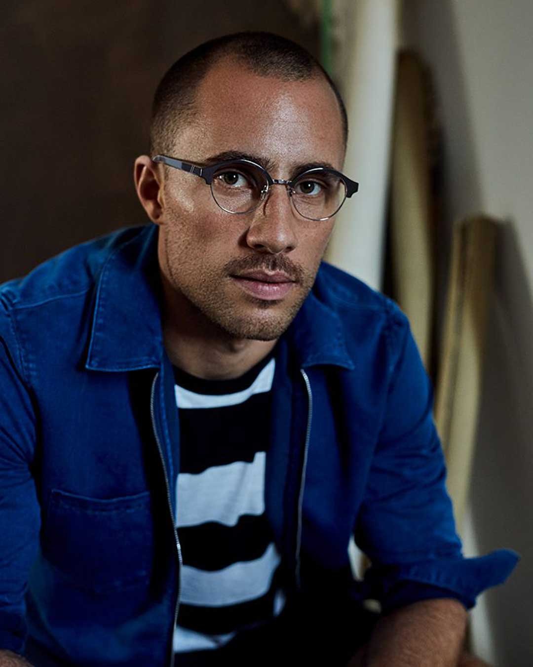 Man with shaved hair wearing blue shirt and Clubmaster spectacle frame in dark room