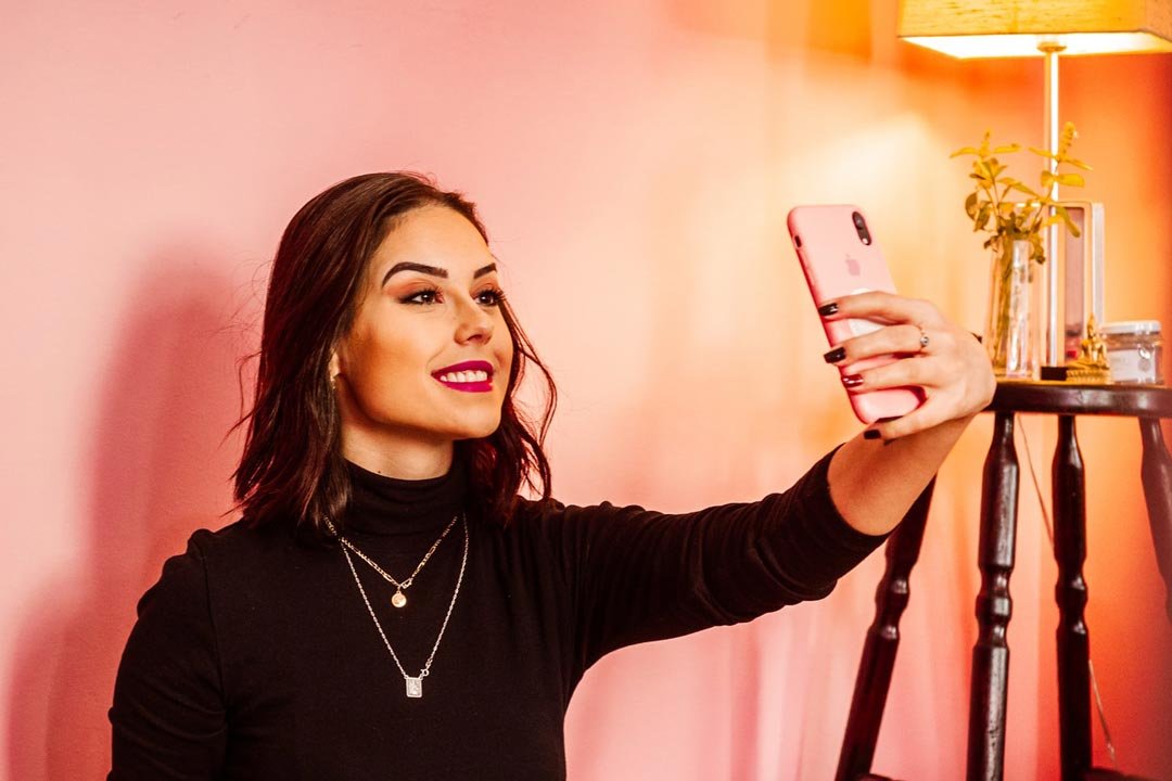 Young woman sitting in front of pink wall smiling taking a photo of herself using her smartphone